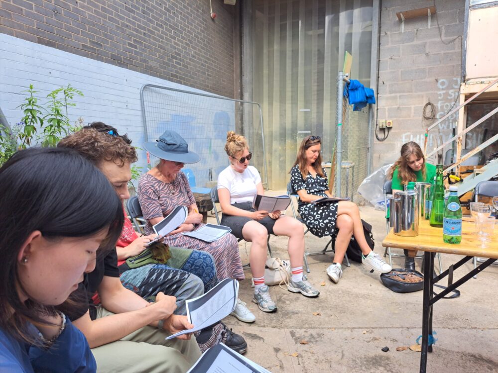 Group of six people sat on chairs in a semi-circle facing downwards and holding notebooks and pens