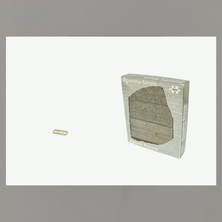 A photograph of a poster on a grey background. On the poster: A white background with a photograph of the box of a 'Rosetta Stone' jigsaw puzzle on the right, and a razorblade on the left.