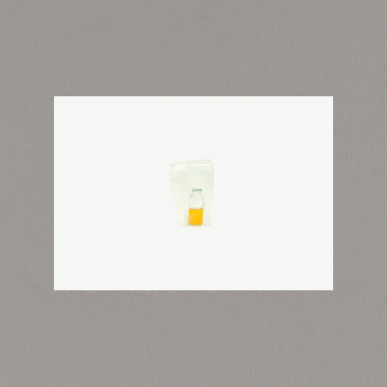 A photograph of a postcard on a grey background. On the postcard: A white background with a photograph of a bottle in a clear plastic bag. There is yellow liquid inside the bottle.