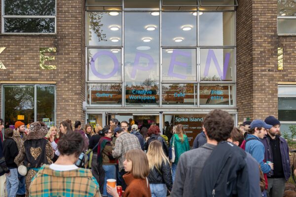 Lots of people standing and chatting outside of a building with the word 'OPEN' written on the windows above them.