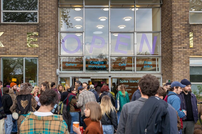 Lots of people standing and chatting outside of a building with the word 'OPEN' written on the windows above them.
