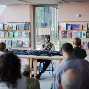 Photo of a Black artist sitting at a DJ desk and speaking into a mic in front of an audience.