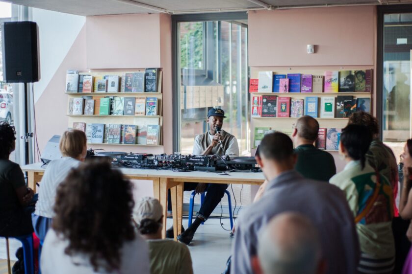 Photo of a Black artist sitting at a DJ desk and speaking into a mic in front of an audience.
